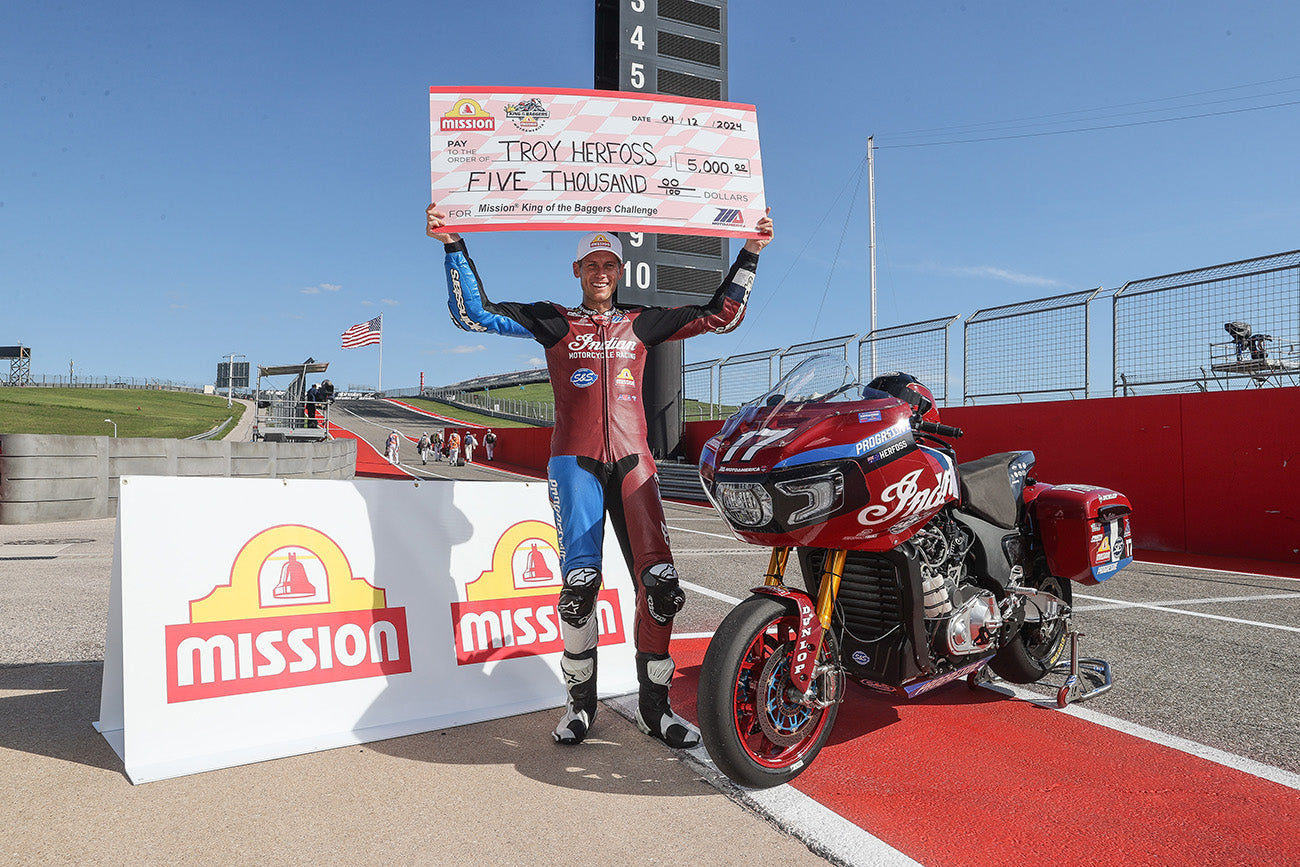 Troy Herfoss triumphantly holding up a $5,000 check after winning the Mission King of the Baggers Challenge at the MotoGP RedBull Grand Prix of the Americas. He is dressed in a full racing suit, standing next to his red Indian motorcycle, with the iconic Circuit of the Americas track in the background under a clear blue sky.