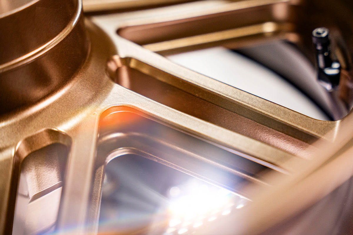 Detailed close-up of a bronze motorcycle wheel by Dymag, highlighting the elegant spoke design illuminated by bright light reflections, creating a warm metallic glow.