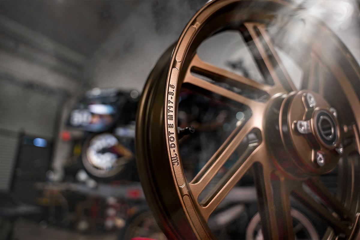 Close-up of a bronze Dymag motorcycle wheel with embossed product details along the rim edge, set against a dark, blurred workshop background.