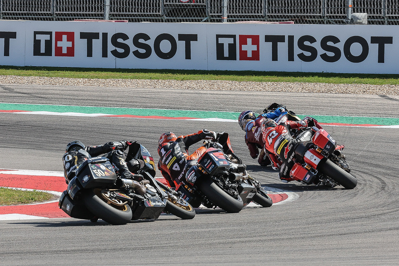 Intense racing action captured at the MotoGP RedBull Grand Prix of the Americas, featuring a tight group of motorcycles leaning dramatically through a sharp turn. The riders, clad in colorful racing suits, are competing fiercely under the watchful eyes of Tissot and Swiss cross banners, emphasizing the high stakes and precision of the race.