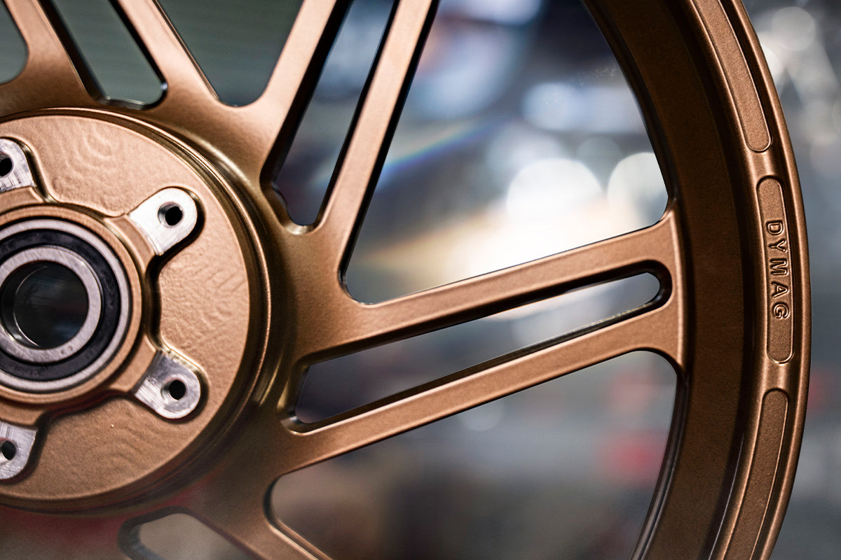 Close-up of a golden Dymag racing wheel with intricate spoke design and hub details, set against a softly focused, metallic workshop background.