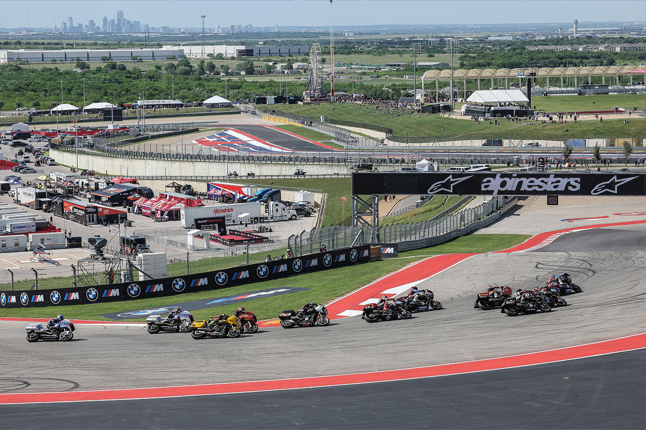 Wide-angle view of the MotoGP race track at the Circuit of the Americas in Austin, Texas, with a group of motorcycles navigating a curve against a panoramic backdrop of the city skyline. The scene captures the bustling pit lane filled with team trailers and spectator areas, while the prominent Alpinestars banners emphasize the racing theme.
