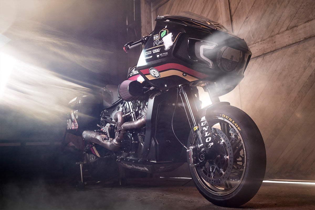 Motorcycle equipped with Dymag wheels in a dramatic workshop setting, highlighted by shafts of light illuminating the dark surroundings and casting a warm glow on the bike.