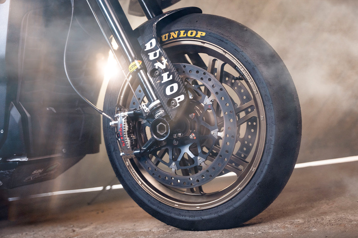 Close-up of a golden Dymag racing wheel with intricate spoke design and hub details, set against a softly focused, metallic workshop background.