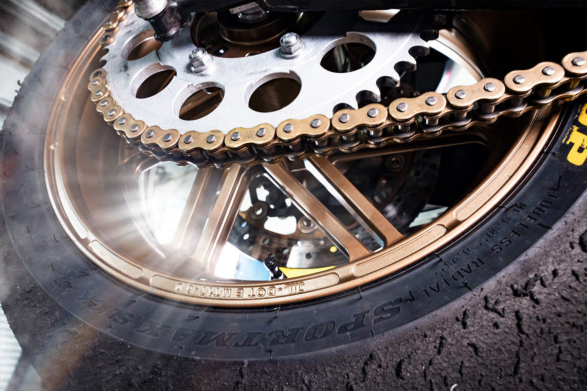 "Intensely detailed image focusing on the bronze Dymag wheel, chain, and sprocket of a motorcycle, emphasizing the gritty texture of the tire and the precision engineering of the components.