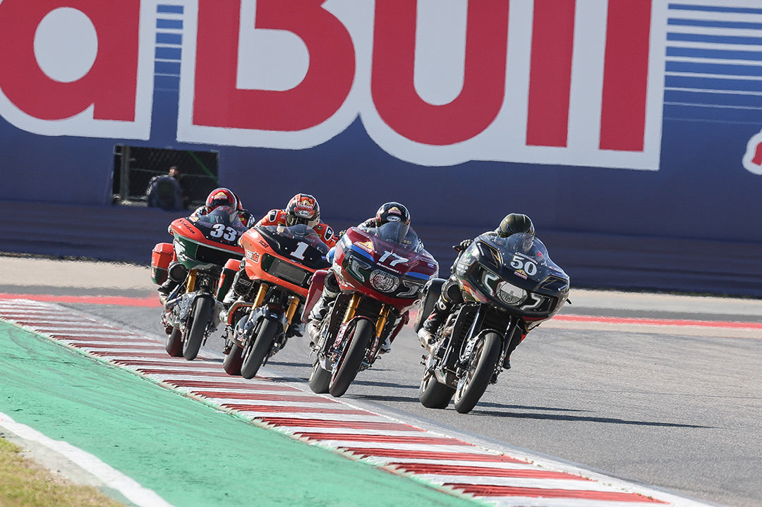 The leading group of motorcycles charges forward at the Circuit of the Americas, with a focus on bikes bearing numbers 33, 1, and 77, illustrating the high stakes and competitive intensity under a large Red Bull logo.
