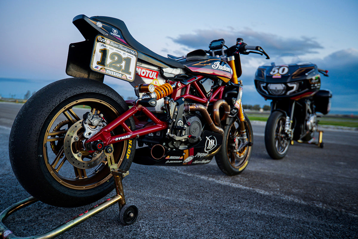 Two racing motorcycles with Dymag wheels parked side by side at sunset, with vibrant sky colors reflecting off the metallic surfaces of the bikes.