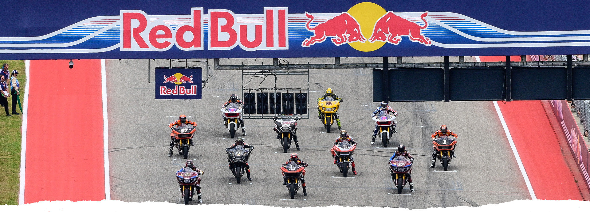 A thrilling start at the MotoGP RedBull Grand Prix of the Americas in Austin, Texas, with a lineup of motorcycle racers speeding off under the massive Red Bull logo arch. The racers, in a mix of vibrant liveries, are captured just as they launch from the grid, with the track bordered by striking red and blue boundaries, capturing the excitement and competitive spirit of the race.