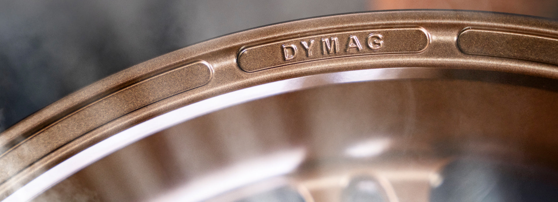 Extreme close-up of the rim of a bronze Dymag racing wheel, showcasing the embossed 'DYMAG' logo and detailed texture of the metal surface, with a blurred dark background enhancing the focus on the wheel's craftsmanship.
