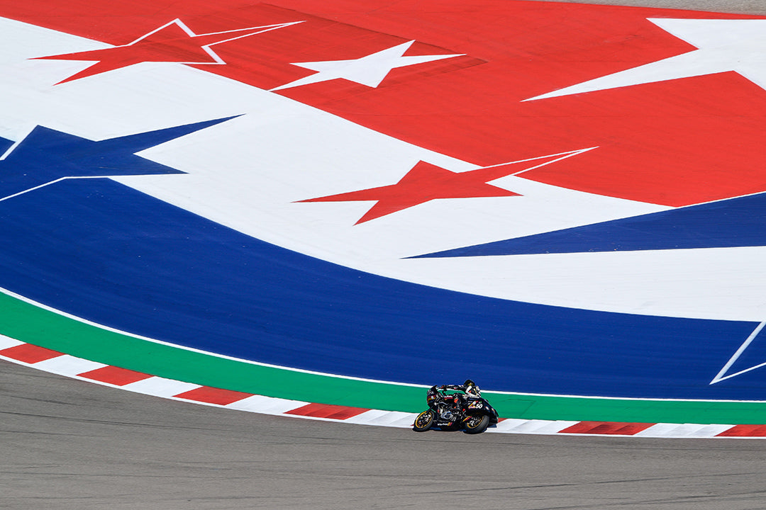 A motorcycle leans into a curve on a visually striking section of the track painted with large white stars on a red and blue background, emphasizing the Americana theme at the Circuit of the Americas during the MotoGP race.
