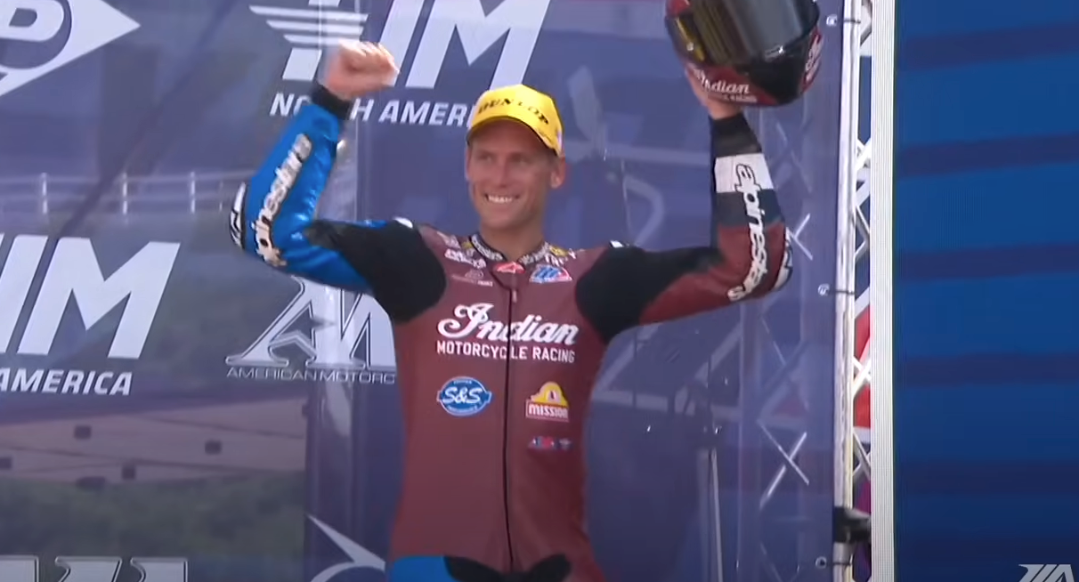 Troy Herfoss celebrates a victory at Michelin Raceway Road Atlanta, raising his helmet in triumph. He's wearing a full Indian Motorcycle racing suit and a bright yellow cap, smiling broadly on the podium under a backdrop featuring the MotoAmerica and AMA Pro Racing logos.